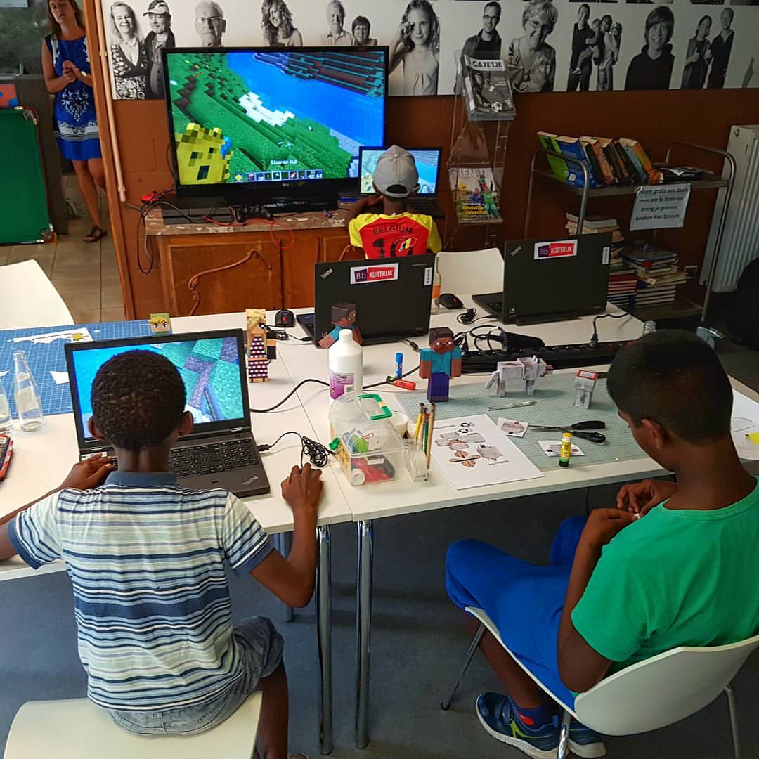 Children playing Minecraft on laptops and a large screen in a library.